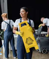 happy bi-racial woman holding caution board near coworkers and floor scrubber machine