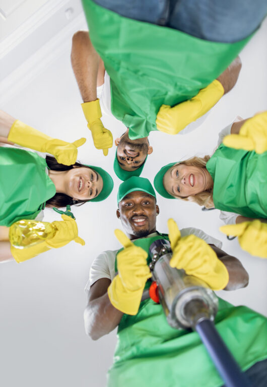 Low angle view of four male and female cleaners in green apron, hat and yellow gloves standing in circle and looking at camera with thumbs up. Multiracial janitors at work.